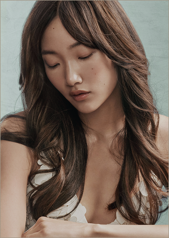 - Ibi David Salon in Westlake, OH A woman with long, wavy brown hair looks downward with a neutral expression, wearing a white top against a light blue background. - Ibi David Salon in Westlake, OH